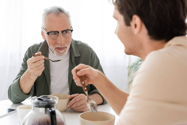 zralý muž v brýlích drží lžíci a při snídani se dívá na rozmazaného syna  - Fotografie, Obrázek