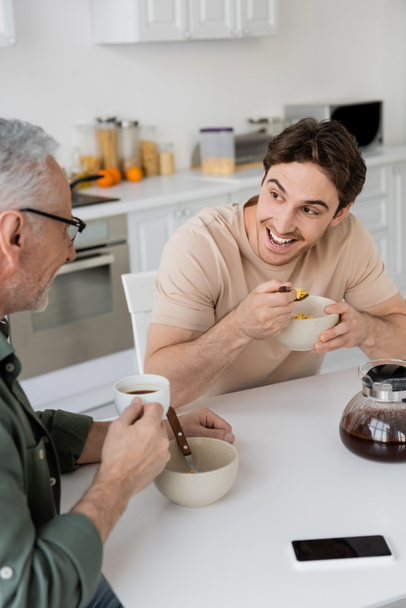 hombre sorprendido escuchando a padre mientras que come hojuelas de maíz cerca de la cafetera en la cocina - Foto, Imagen