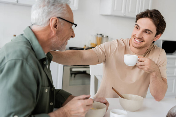 cheerful young man holding coffee cup and touching shoulder of dad while talking to him during breakfast - Photo, Image