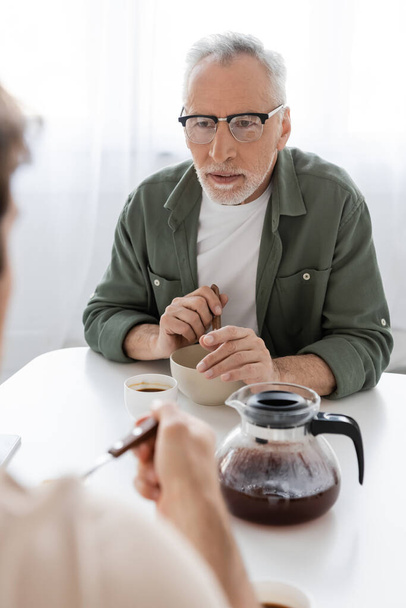 uważny człowiek w okularach patrząc na rozmazanego syna w pobliżu śniadania i dzbanek do kawy na stole w kuchni - Zdjęcie, obraz
