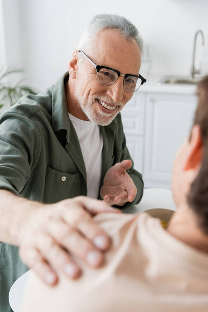 smiling man in eyeglasses pointing with hand and touching shoulder of blurred son during conversation in kitchen - Photo, image