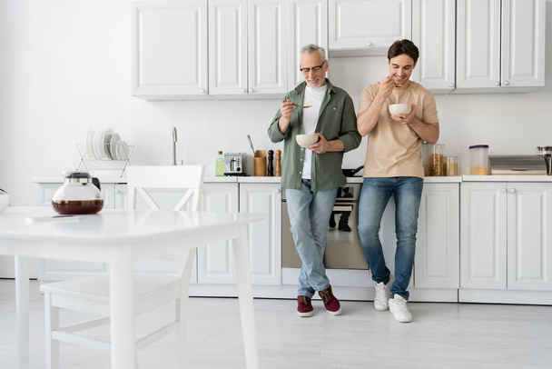 full length of mature father and young son having breakfast while standing in modern kitchen  - Zdjęcie, obraz