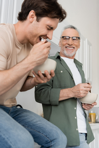 happy man in eyeglasses looking at young son having breakfast and laughing on blurred foreground - Fotoğraf, Görsel