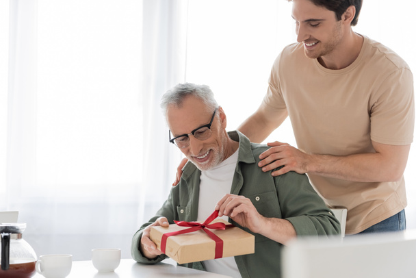 smiling guy touching shoulders of pleased dad opening fathers day present - Photo, Image