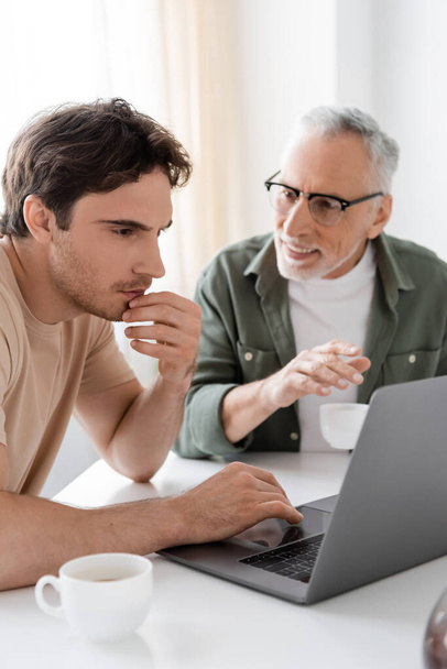 Mutfaktaki düşünceli oğlunun yanında, elinde kahve fincanı olan beyaz saçlı bir adam dizüstü bilgisayarı işaret ediyor. - Fotoğraf, Görsel