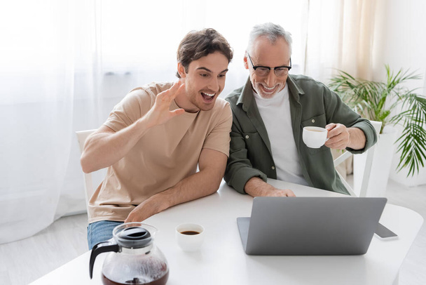 興奮した男手を振ってビデオ通話中にノートパソコンで明るいお父さんと一緒にコーヒーカップ - 写真・画像