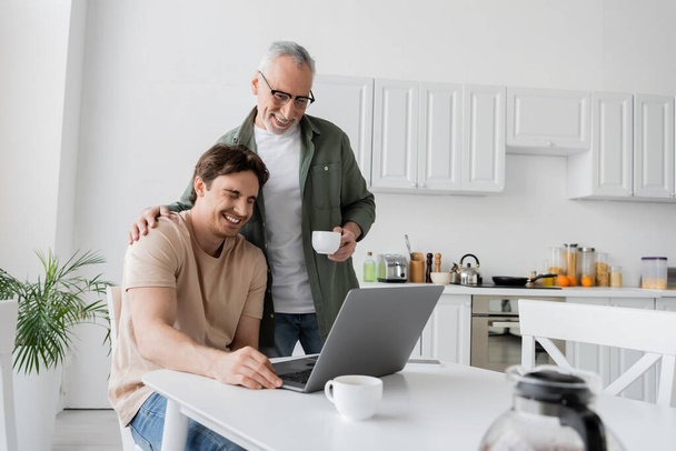 érett és mosolygós férfi kávéscsésze megható váll fia nevetés mellett laptop a konyhában - Fotó, kép