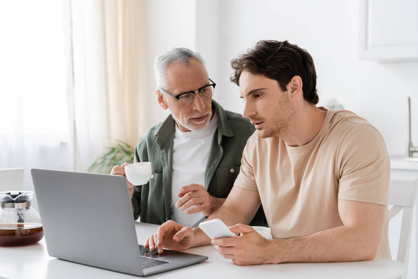 Gözlüklü, elinde kahve fincanı olan gri saçlı bir adam ve yetişkin oğlunun yanındaki dizüstü bilgisayarı işaret ediyor. - Fotoğraf, Görsel