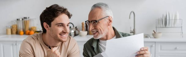 mature man in eyeglasses showing documents to happy son, banner - Фото, изображение