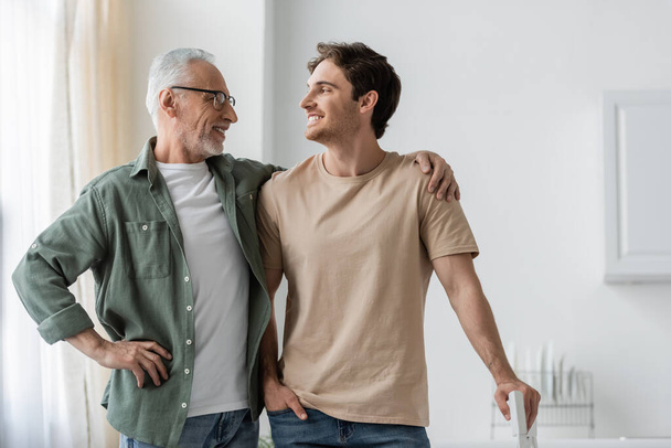 cheerful grey haired man and young son looking at each other while posing in kitchen - Photo, Image