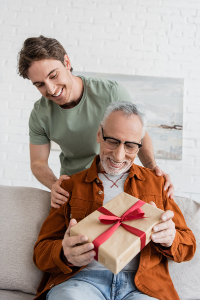 cheerful man hugging shoulders of happy dad holding fathers day present on couch at home - Photo, Image