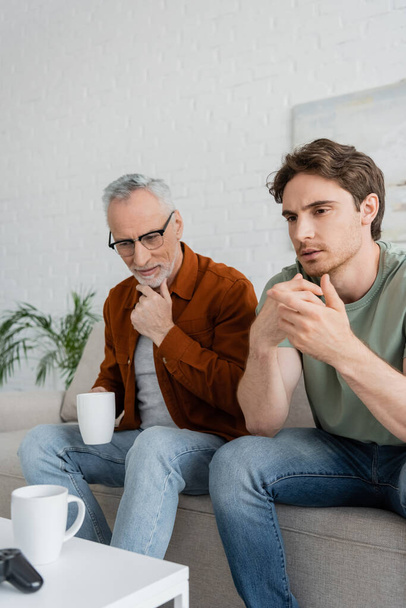 thoughtful mature man with tea cup touching beard while sitting near serious son  - Фото, зображення