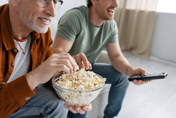 bijgesneden weergave van glimlachende senior man met kom van popcorn kijken tv met opgewonden zoon op wazig achtergrond - Foto, afbeelding