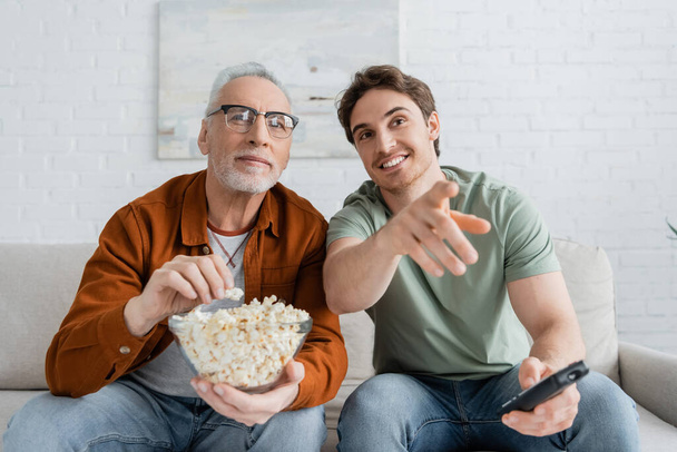 sonriente hombre señalando con el dedo mientras ve la televisión con el padre sosteniendo un tazón de palomitas de maíz - Foto, Imagen
