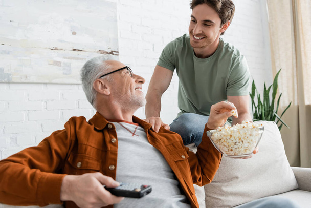 cheerful guy holding bowl of popcorn near dad sitting on couch with remote controller - Zdjęcie, obraz