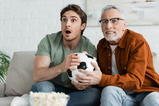 thrilled guy with tense dad holding soccer ball while watching football championship on tv - Photo, Image