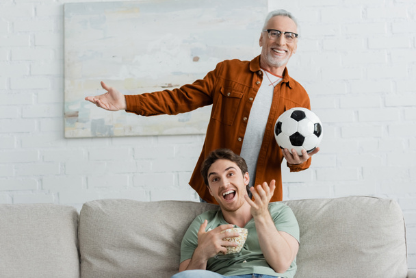 happy man with soccer ball and cheerful son with bowl of popcorn gesturing while watching football match on tv - Photo, Image