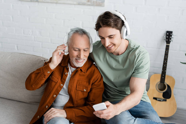 sonriente hombre mostrando teléfono inteligente a feliz hombre de pelo gris en auriculares inalámbricos mientras escucha música en casa - Foto, imagen