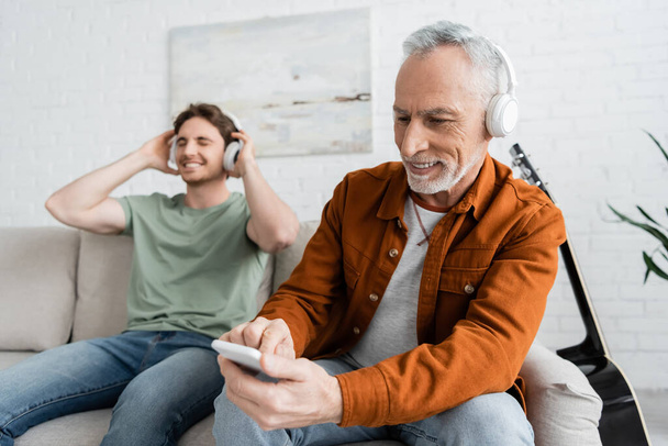 homme mature et heureux dans les écouteurs sans fil en utilisant un smartphone près de son écoute de musique sur fond flou - Photo, image