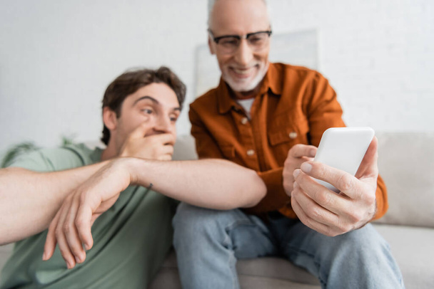 happy and mature father with beard holding smartphone near young son on blurred background  - Photo, image