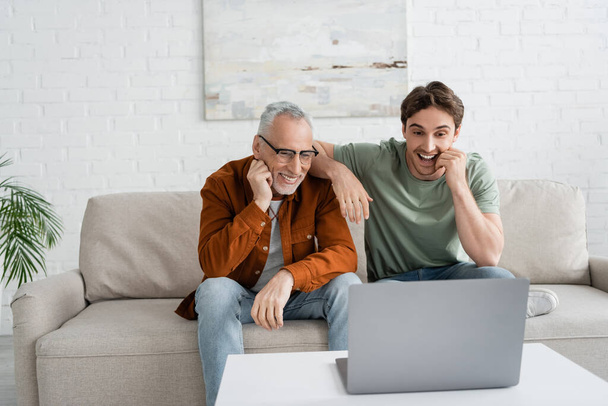 fröhlicher junger Mann schaut Komödienfilm auf Laptop mit reifem Vater in Brille  - Foto, Bild