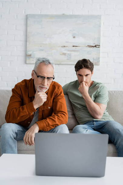 眼鏡をかけた大人の父親とラップトップで映画を見ている深刻な若い男  - 写真・画像