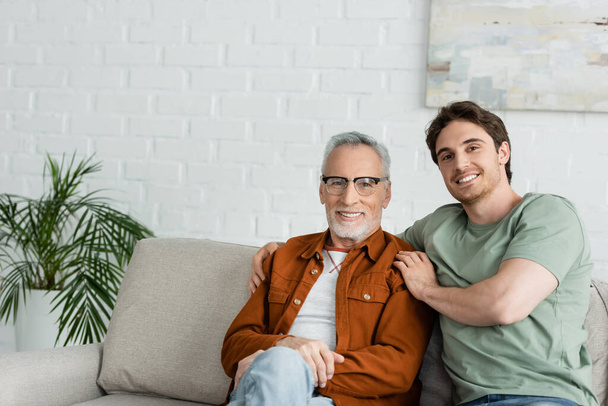 щасливий молодий чоловік обіймає зрілого батька в окулярах, сидячи на дивані
 - Фото, зображення