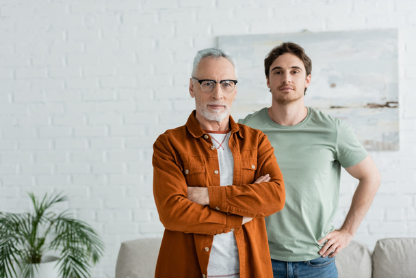 young man standing with mature father in eyeglasses posing with crossed arms at home - Fotoğraf, Görsel