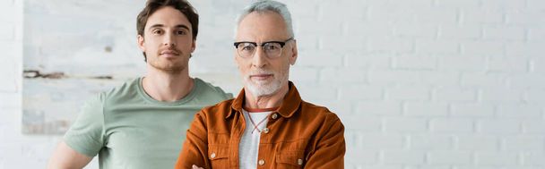 young man standing with mature father in eyeglasses at home, banner - Фото, изображение