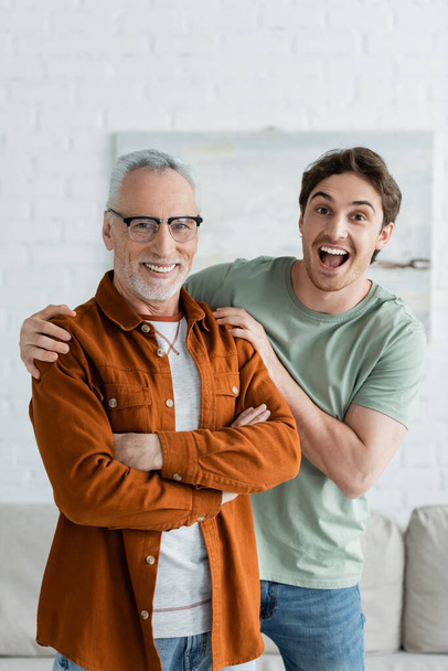 amazed young man hugging mature father in eyeglasses standing with crossed arms at home - Photo, Image