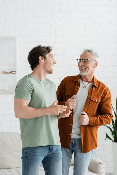 Ευτυχισμένος νεαρός άνδρας που μιλάει με χαμογελαστό ώριμο πατέρα με γυαλιά στο σπίτι - Φωτογραφία, εικόνα