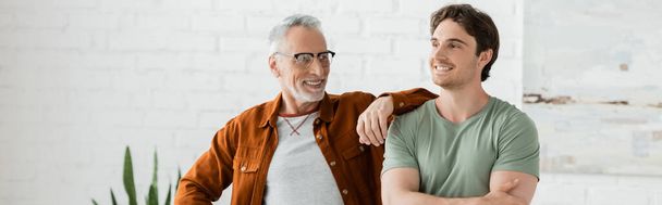 happy mature father in eyeglasses leaning on shoulder of cheerful young son posing with crossed arms at home, banner - Photo, Image