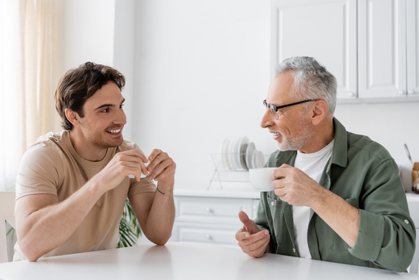 felice giovane che beve caffè del mattino con sorridente e maturo padre a casa - Foto, immagini