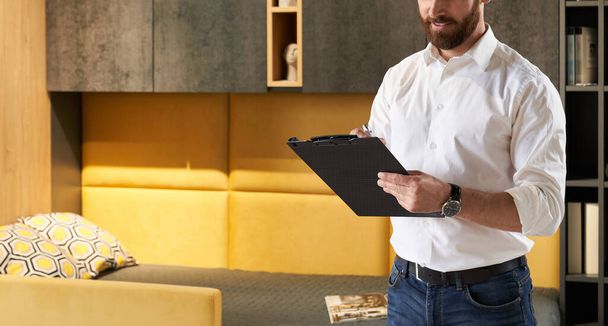 Unrecognizable bearded male retailer making notes, while working in furniture store. Crop view of sales manager writing in tablet, while standing in front of bedroom furniture. Concept of workplace.  - 写真・画像
