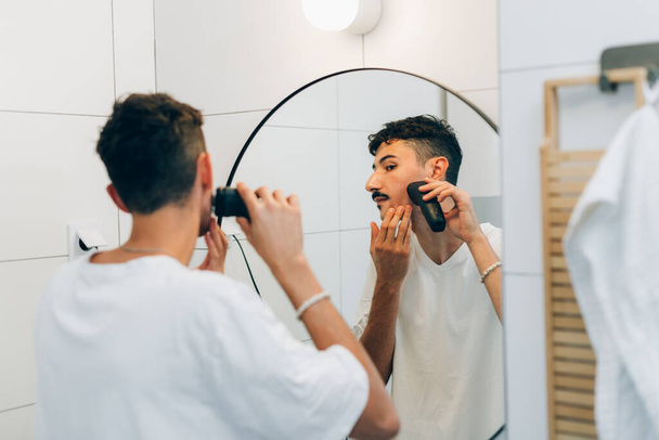 Young man shaves his beard with a trimmer - Fotó, kép