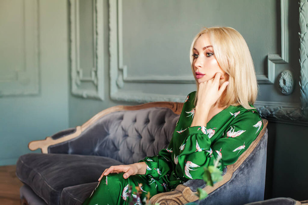 Belle femme blonde souriante et heureuse en robe verte assise sur un canapé en velours - Photo, image