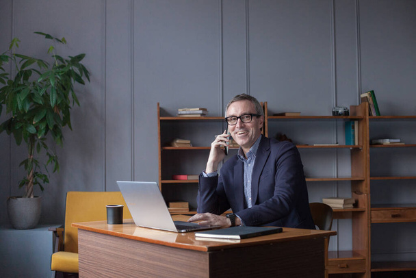 Šťastný podnikatel drží svůj mobilní telefon v domácí kanceláři - Fotografie, Obrázek