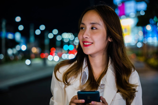 Business woman using mobile phone walking through night city street while waiting car to pick up home, Beautiful young smiling female texting work message on smartphone outside office, social media - Foto, imagen