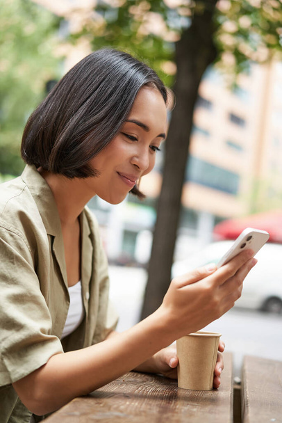 Primer plano retrato de chica asiática sentado en la cafetería al aire libre, beber café y el uso de teléfonos inteligentes, la lectura de teléfono móvil. - Foto, Imagen