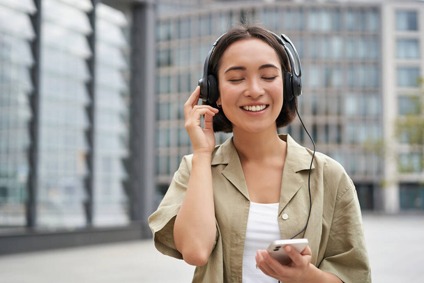 Koncepcja usług strumieniowych. Szczęśliwa azjatycka dziewczyna słucha muzyki w słuchawkach, trzyma telefon komórkowy, wybierając utwór lub podcast, chodzenie po ulicy. - Zdjęcie, obraz