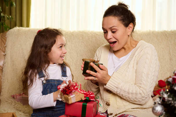 Heerlijke liefhebbende moeder verheugt zich terwijl ze een kerstcadeau ontvangt van haar schattige dochtertje. Mooie baby in blauwe denim overalls geeft een schattig kerstcadeau doos aan haar zorgzame moeder. Gelukkig Nieuwjaar - Foto, afbeelding