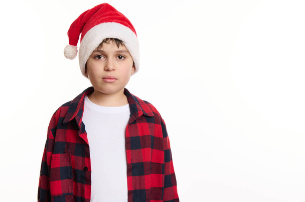 Isolated portrait on white background of a handsome Caucasian preteen boy in Santa hat, in anticipation of Christmas holidays. Pure joy. Winter spirit. New Year party. Copy space for advertising text - Photo, image