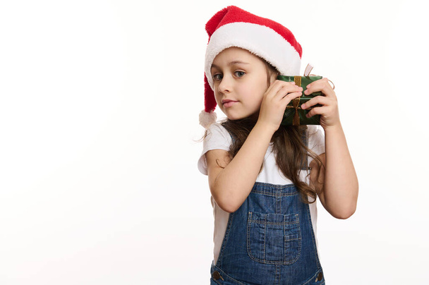 Retrato aislado sobre fondo blanco de una niña feliz, adorable y curiosa en sombrero de Santa, sacudiendo un regalo de Navidad y adivinando lo que hay dentro. Copia el espacio publicitario. Hora de abrir cajas de regalo de Navidad - Foto, Imagen