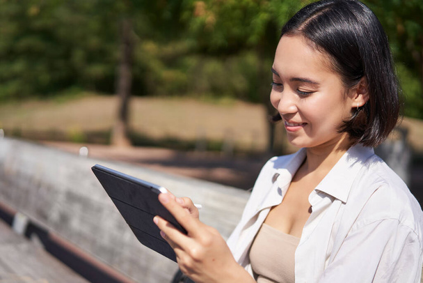 Ritratto di giovane donna asiatica che attinge aria fresca nel parco, seduta con tablet grafico e penna digitale, sorridente felice. - Foto, immagini