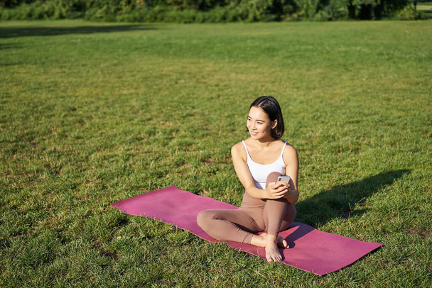 Femme dans le parc, regarder la vidéo de yoga sur smartphone, méditer sur l'air frais, assis sur un tapis en caoutchouc. - Photo, image