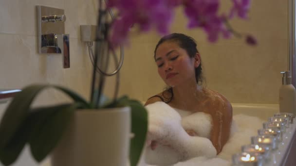 Hermosa joven asiática disfrutando de tomar un baño de burbujas en un ambiente relajante. Relajación y tratamiento de spa en el baño en casa para el final del día. Señora tratando a sí misma con baño lleno de espuma. - Metraje, vídeo