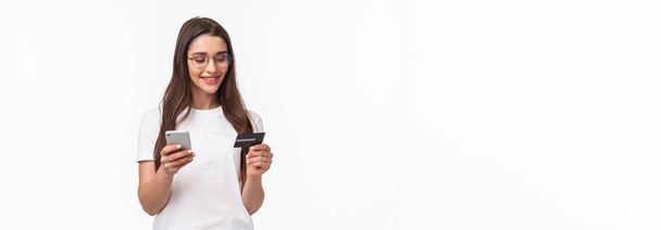 Πορτρέτο της ελκυστική νεαρή γυναίκα σε γυαλιά που κατέχουν πιστωτική κάρτα και smartphone, εισάγετε τους αριθμούς ψηφίων του τραπεζικού λογαριασμού της στην εφαρμογή αγορών, online παραγγελία, σταθεί λευκό φόντο. - Φωτογραφία, εικόνα