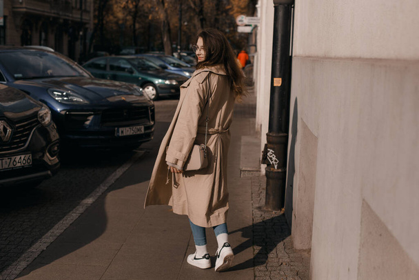 Young woman in a coat outside. Outdoor autumn portrait of young elegant fashionable woman wearing trendy glasses, camel color coat, shoulder bag, walking in street of European city. Copy space - Foto, imagen
