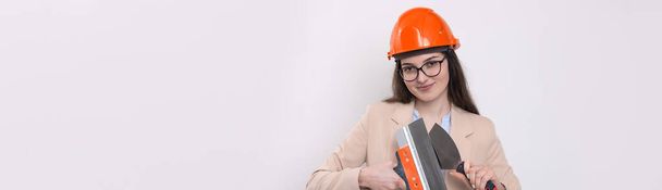 Girl engineer in an orange construction helmet with plastering painting tools in her hands on a white background. - Zdjęcie, obraz