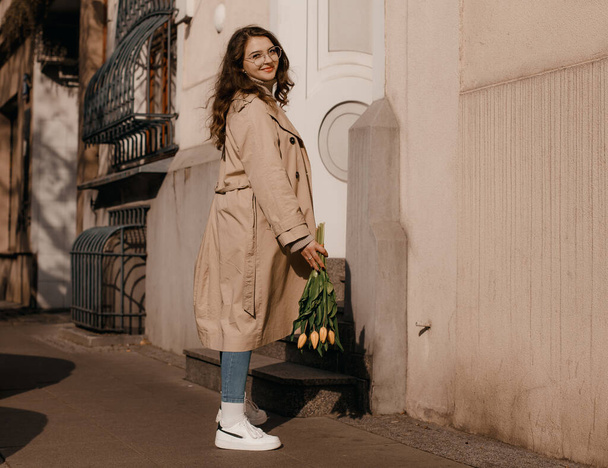 Young woman in a coat outside. Outdoor autumn portrait of young elegant fashionable woman wearing trendy glasses, camel color coat, shoulder bag, walking in street of European city. Copy space - Photo, Image